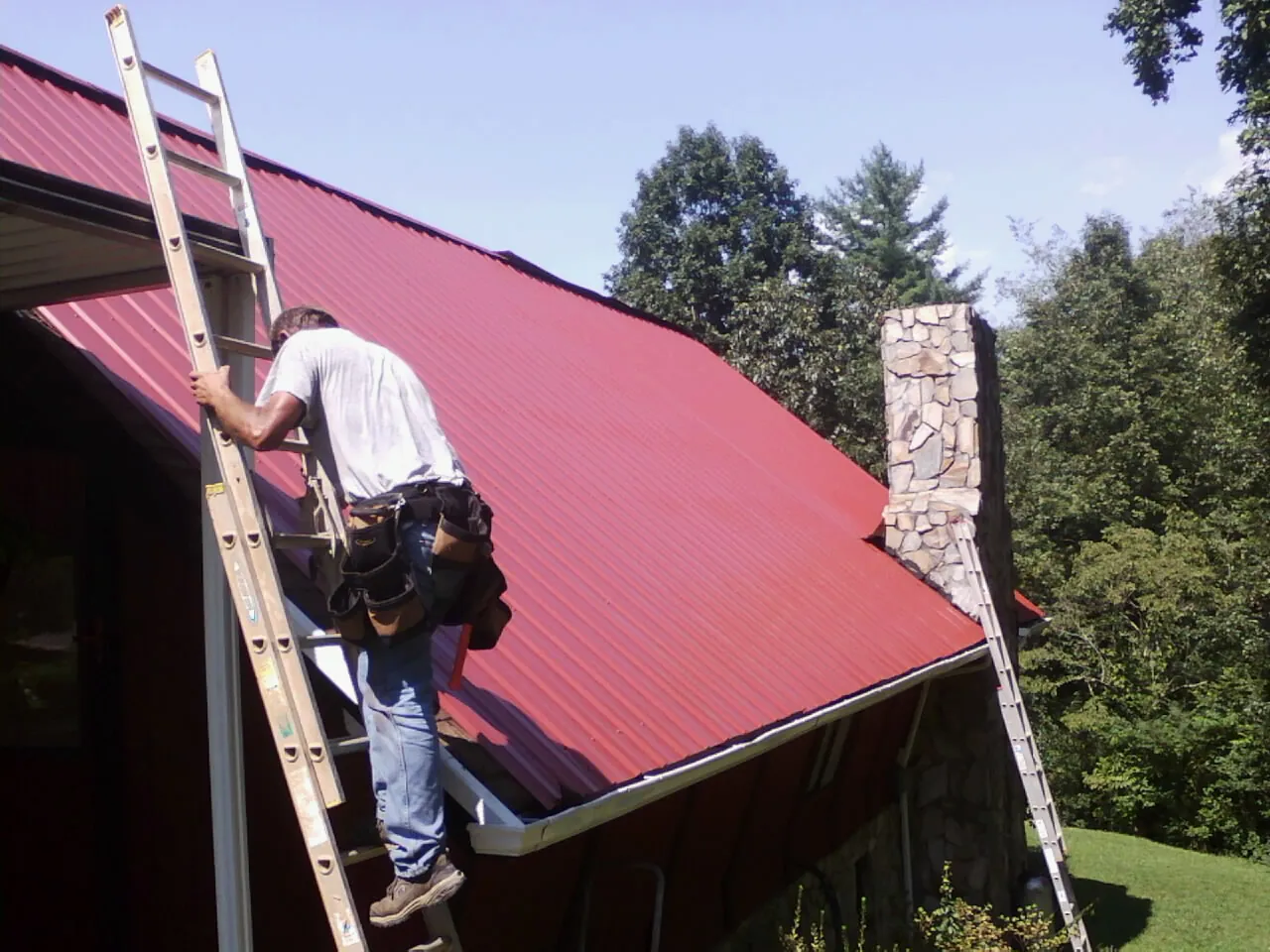 Roofer on a ladder beside a red metal roof and stone chimney — Soffit & Fascia Repair by Roofing Brothers in Biddeford